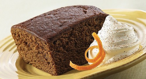 Square of gingerbread on a plate, garnished with a swirl of whipped cream and a curled strip of orange peel