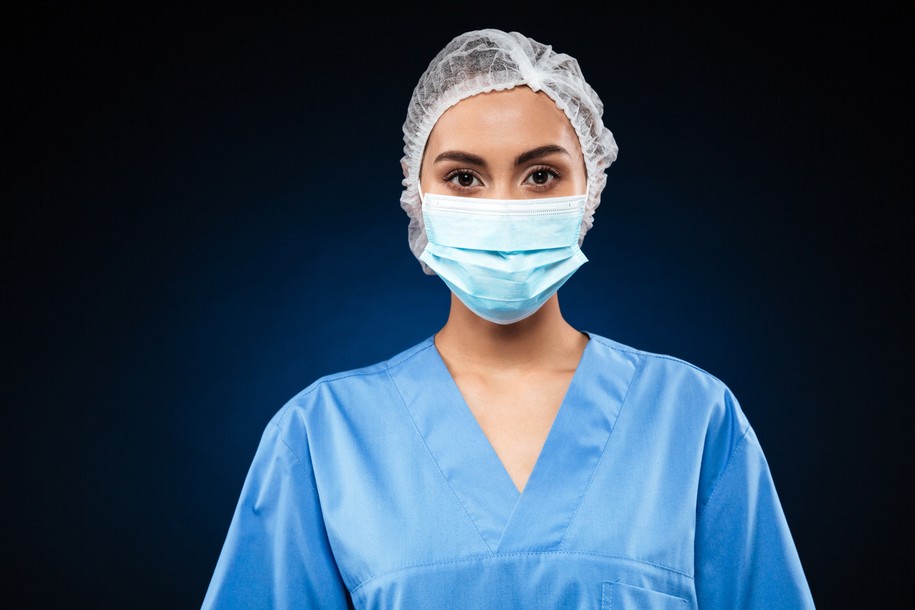 Young doctor in medical mask and cap wearing blue uniform and looking camera isolated over black